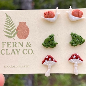 Könnte beinhalten: Drei Paar Ohrstecker auf einer Präsentationskarte mit der Aufschrift Fern & Clay Co. 14k Gold Plated. Die Ohrringe zeigen Miniaturfiguren aus Ton: weiße Schnecken mit orangefarbenen Häusern, grüne Frösche und rot-weiße Pilze mit Punkten.
