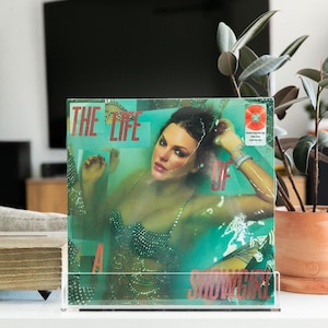 Può includere: Un album in vinile intitolato "The Life Of A Showgirl" con una donna con un top scintillante immersa nell'acqua. La copertina dell'album è esposta in un supporto in acrilico trasparente su una superficie bianca.