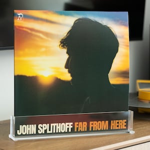 Può includere: Una copertina di un album in vinile con la silhouette di una persona sullo sfondo di un tramonto. Il titolo dell'album "FAR FROM HERE" e il nome dell'artista "JOHN SPLITHOFF" sono visualizzati su un supporto in acrilico trasparente.