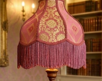Op de afbeelding: Een vintage vloerlamp met een roze lampenkap met een gouden damastpatroon en franjes. De lamp heeft een houten voet en staat in een kamer met een klassiek, elegant decor.