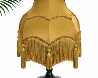 May include: A mustard yellow fabric lampshade with a scalloped edge, decorative braided trim, and long hanging fringe tassels. The shade is mounted on a solid black circular base, creating a structured silhouette with a smooth, matte finish.