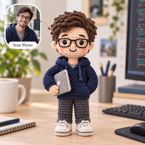 May include: A handmade crochet doll featuring a person with brown hair, black-rimmed glasses, and a navy blue hooded sweatshirt. The doll holds a small gray rectangular object and wears dark gray pants with white shoes, inspired by a photo.