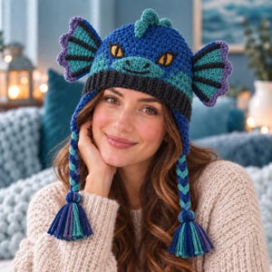 May include: A crocheted dragon hat in shades of blue, teal, and purple. The hat features a dark blue ribbed brim, two large side fins, a central crest, and two long braided earflaps ending in multi-colored tassels. The dragon face has yellow eyes.
