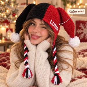 May include: A two-toned black and red crocheted jester hat featuring two long ear flaps with white pom-poms at the ends and braided tassels in red, black, and white. The red side of the hat displays the white embroidered text LUNA.