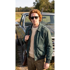 May include: A dark green suede bomber jacket with a front zipper, ribbed cuffs, and two flap pockets, worn over a cream-colored cable knit sweater. The outfit includes dark sunglasses and a brown leather strap slung over the shoulder.