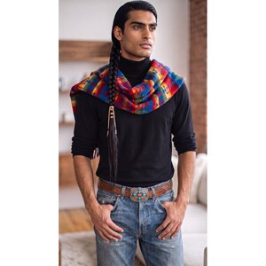May include: A black turtleneck sweater paired with blue denim jeans and a brown leather belt featuring a silver-toned buckle with turquoise and coral inlays. A multi-colored patterned blanket scarf with red, blue, and yellow geometric designs is draped.