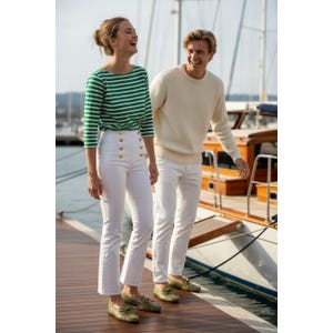 May include: A woman wearing a green and white striped long-sleeve shirt, white pants with gold buttons, and gold loafers. A man wears a cream-colored sweater, white pants, and gold loafers. They are standing on a wooden dock near a boat.