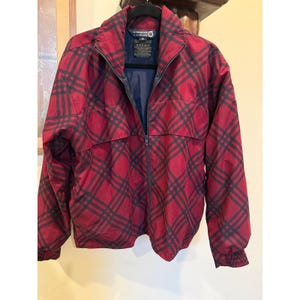 Puede incluir: Una chaqueta cortavientos de cuadros rojos y negros con cremallera frontal completa, cuello alto y mangas largas con puños elásticos. La etiqueta interior muestra el texto Sunderland of Scotland con una indicación de talla M e instrucciones.