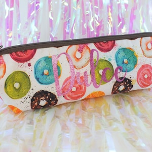 May include: A rectangular pencil case with a brown zipper and a colorful donut pattern. The case features donuts in various colors, including pink, blue, green, and brown, with the name "Dulce" in pink glitter.