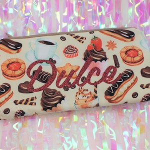 May include: A rectangular beige pencil case with a zipper closure, adorned with a pattern of various sweets like eclairs, cupcakes, and cookies. The word "Dulce" is written in red glitter.