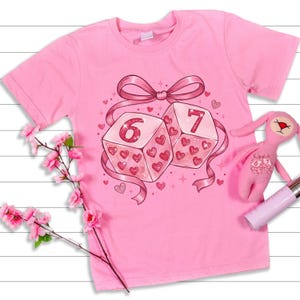 May include: A pink t-shirt with a graphic of two dice, showing the numbers 6 and 7, adorned with hearts and a pink ribbon. A pink toy bunny and a pink flower branch are also visible.