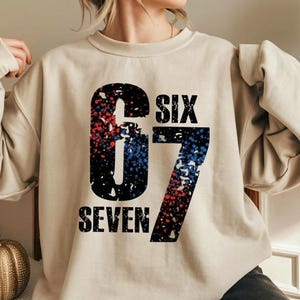 May include: Beige sweatshirt featuring the text "SIX 67 SEVEN" in a distressed font. The numbers and text are black with red and blue splatter effects. The sweatshirt is a neutral colour.