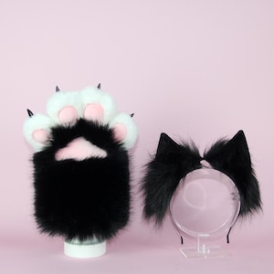 May include: A pair of black and white faux fur cat paw gloves with pink paw pads and black claws, and a black cat ear headband with fluffy fur, both on a pink background.