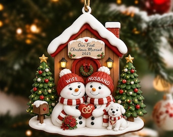 May include: A house-shaped Christmas ornament featuring two snowmen wearing red hats labeled Wife and Husband, holding a red heart with gold rings. The ornament includes two green Christmas trees, a small white dog, and a sign reading Our First Christmas Married 2025.