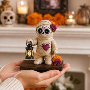 May include: A mummy figurine wrapped in beige bandages with a sugar skull face featuring blue and red floral patterns. The figure holds a small black lantern with a glowing light and has a purple heart on its chest, adorned with orange and purple flowers.