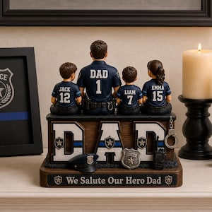 May include: A decorative figurine featuring a police officer figure with three children, each wearing a jersey with a number. The base reads "DAD" and "We Salute Our Hero Dad". A framed police badge and candles are also present.
