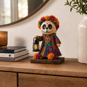 May include: A decorative figurine of a skull-faced character with a floral crown, wearing a purple and teal patterned dress with a heart motif. The figure holds a small black lantern with a glowing light and stands on a rectangular wooden base.