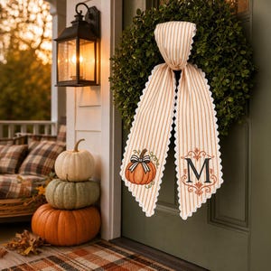 May include: A green boxwood wreath hanging on a door, adorned with a long, cream and orange striped fabric ribbon. The ribbon features a scalloped white edge, an embroidered orange pumpkin with a black bow, and a large black letter M monogram.