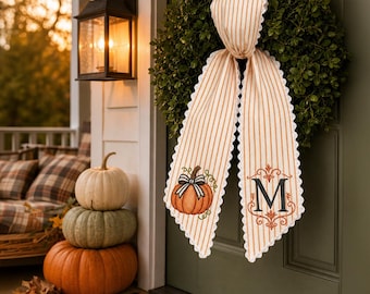May include: A green boxwood wreath hanging on a door, adorned with a long, cream and orange striped fabric ribbon. The ribbon features a scalloped white edge, an embroidered orange pumpkin with a black bow, and a large black letter M monogram.
