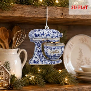 May include: A flat, hanging ornament shaped like a stand mixer, featuring a white and blue pattern of reindeer, sleighs, and holiday motifs. The mixing bowl is decorated with a windmill design and the name Sophia in gold script lettering.