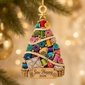 Op de afbeelding: Een driehoekig kerstboomornament gemaakt van kleurrijke garenklosjes, stofrollen, knopen en een meetlint. De basis bevat een houten plaatje met de tekst Sew Happy 2026 en een icoon van een klosje, bekroond met een gouden ster en een ophanglus.