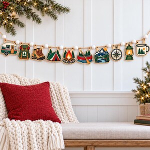 May include: A decorative garland featuring ten wooden cutouts suspended on a string with white beads. The shapes include a camper, backpack, hiking boots, tent, campfire, canoe, mountain landscape, compass, lantern, and a coffee mug.
