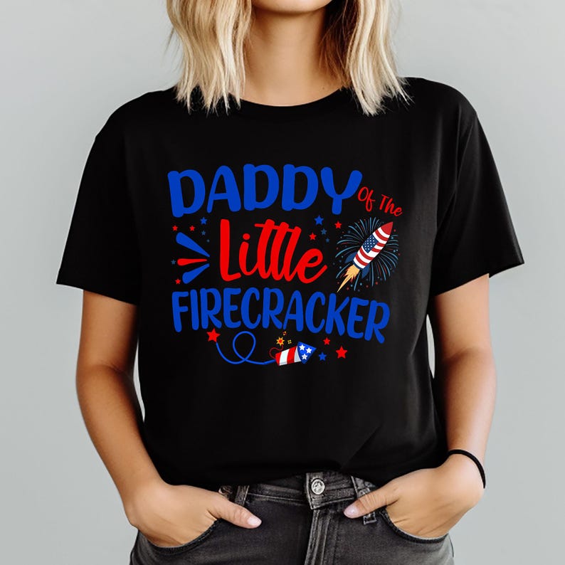 May include: Black t-shirt with the text "DADDY of The Little Firecracker" in blue and red. The design includes a firework rocket, stars, and a small flag, perfect for celebrating Independence Day.