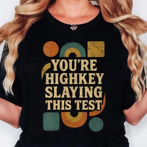 May include: Black t-shirt with the text "YOU'RE HIGHKEY SLAYING THIS TEST" in a retro font. The design includes geometric shapes in orange, teal, and yellow. A necklace with a butterfly pendant is visible.