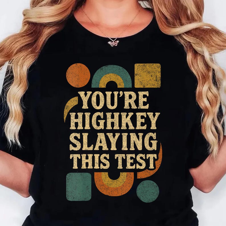 May include: Black t-shirt with the text "YOU'RE HIGHKEY SLAYING THIS TEST" in a retro font. The design includes geometric shapes in orange, teal, and yellow. A necklace with a butterfly pendant is visible.