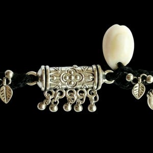 May include: An ornate silver-toned anklet with a black cord. The anklet features a central cylindrical charm with a floral design, dangling bells, and leaf-shaped charms. A white, oval-shaped bead is also attached to the cord. The background is black.