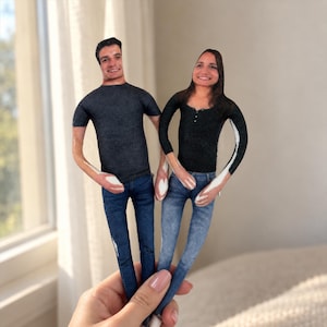 May include: Two custom fabric dolls featuring printed photographic faces of a person with short dark hair in a grey t-shirt and blue jeans, and a person with long dark hair in a black long-sleeve shirt and light blue jeans, held together by the base.