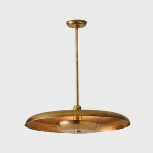 May include: A brass-colored pendant light fixture with a disc-shaped shade and a smaller, rounded light source below. The fixture hangs from a slender rod attached to a circular ceiling mount. The overall design is minimalist and modern.