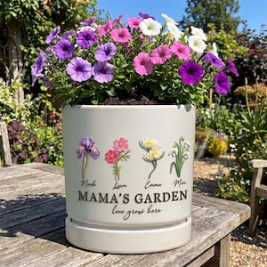 May include: A cream-colored ceramic planter filled with vibrant purple, pink, and white petunias. The planter is decorated with floral illustrations and the text "MAMA'S GARDEN love grows here" along with names. The planter sits on a wooden table.