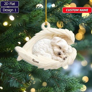 May include: A flat, white, custom-name Christmas ornament featuring a sleeping white puppy nestled within a pair of detailed angel wings. The ornament includes a gold hanging string and the word Name printed in black script on the lower wing.