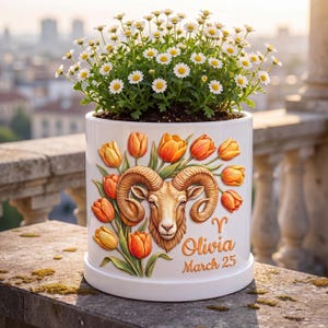 Puede incluir: Una maceta de cerámica blanca con un diseño de cabeza de carnero y tulipanes naranjas y amarillos, llena de margaritas. La maceta tiene el nombre "Olivia" y la fecha "25 de marzo" impresos. La maceta está sobre una repisa de piedra.