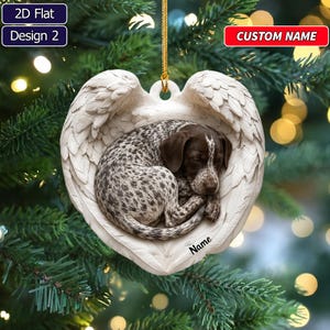 May include: A heart-shaped Christmas ornament featuring a sleeping brown and white spotted puppy nestled within a pair of white angel wings. The ornament includes a gold hanging string and the text Name printed in black on the lower right side.