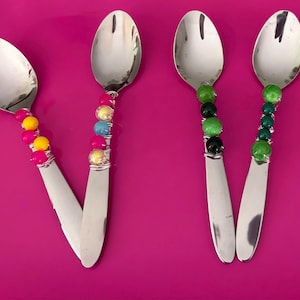 May include: Six stainless steel spoons with decorative handles. Each handle is adorned with colorful beads in various combinations of pink, yellow, blue, green, black, white, and red. The spoons are arranged on a bright pink surface.