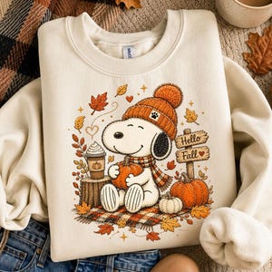 May include: A cream-colored crewneck sweatshirt featuring a graphic of a cartoon dog wearing an orange knit hat and scarf, holding a pumpkin. The design includes a coffee cup, falling autumn leaves, and a wooden sign that reads Hello Fall.
