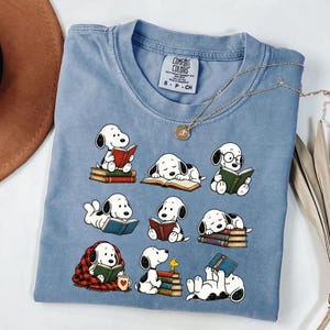 May include: A light blue t-shirt featuring a graphic print of the cartoon character Snoopy in various poses reading books. The shirt has a Comfort Colors brand label at the neck, and a gold-toned chain necklace rests on the fabric surface.