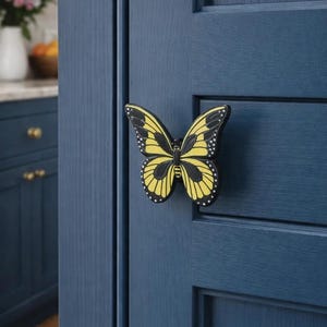 May include: A yellow and black butterfly-shaped cabinet knob is mounted on a dark blue cabinet door. The butterfly has detailed wing patterns and small white dots. The door has a paneled design, and the background shows a glimpse of a kitchen.