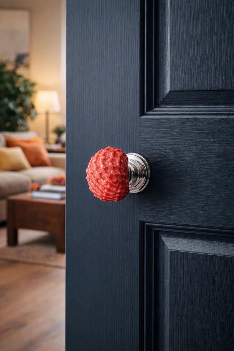 Może przedstawiać: Czerwona, teksturowana gałka do drzwi ze srebrną podstawą zamontowana na ciemnoniebieskich drzwiach. Drzwi są częściowo otwarte, ukazując fragment salonu z sofą i drewnianym stolikiem kawowym. Gałka ma unikalny, dekoracyjny design.
