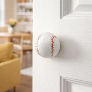 Puede incluir: Un pomo de puerta blanco con diseño de pelota de béisbol y costuras rojas. El pomo redondo está montado en una puerta blanca. El fondo está borroso, mostrando un vistazo de un espacio habitable.