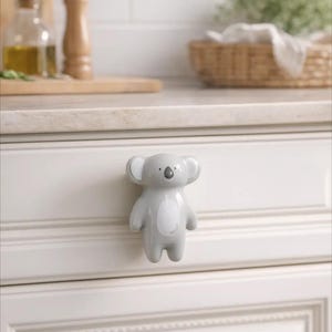 Puede incluir: Un pomo de armario de cerámica gris con forma de koala. El pomo está fijado a una puerta de armario blanca. El koala tiene la barriga y el interior de las orejas blancos, con una pequeña nariz negra. El fondo incluye una encimera de cocina.
