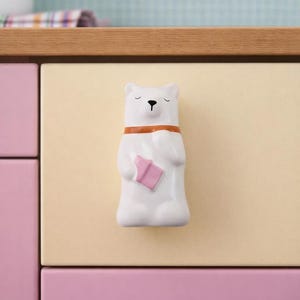 Op de afbeelding: Witte keramische kastknop in de vorm van een beer, met een bruine kraag en een roze boek. De knop is gemonteerd op een lichtgele lade, met daaronder een roze lade. Achtergrond: houten aanrecht en lichtblauwe muur.