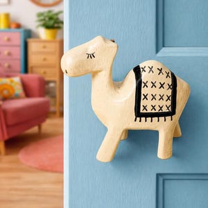 Puede incluir: Un pomo de puerta con forma de camello de color crema con un diseño de silla de montar negro. El pomo decorativo está montado en una puerta azul claro. El camello tiene marcas negras para los ojos y las pestañas.