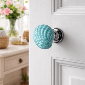 Może przedstawiać: Zbliżenie białych drzwi z dekoracyjną, jasnoniebieską ceramiczną gałką. Gałka ma muszlowy wz&oacute;r i jest zamontowana na chromowanej podstawie. Drzwi są częścią pokoju z rozmytym tłem.