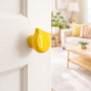 May include: A bright yellow ceramic door knob in the shape of a tulip is mounted on a white door. The knob has a glossy finish and is the focal point of the image. The background shows a living room with a couch and coffee table.