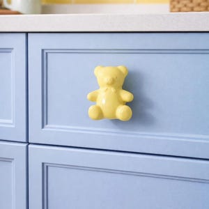Könnte beinhalten: Ein gelber schrankknopf in Teddybärform ist an einer hellblauen Schranktür angebracht. Der Knopf hat eine glänzende Oberfläche und ein kleines Fliegen detail. Der Schrank ist Teil einer Küchenumgebung mit gelbem Fliesenspiegel.