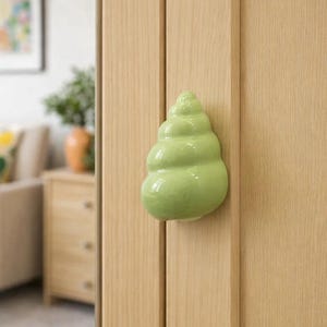 以下が含まれることがあります： ライトグリーンのセラミック製キャビネットノブ、らせん状の貝殻のデザイン。明るい木製のキャビネットドアに取り付けられています。ノブは光沢仕上げで、装飾的なアクセントです。