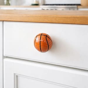 May include: A basketball-themed cabinet knob. The knob is round, with an orange and black design mimicking a basketball. It is mounted on a white cabinet door. The background includes a wooden countertop and a kitchen setting.